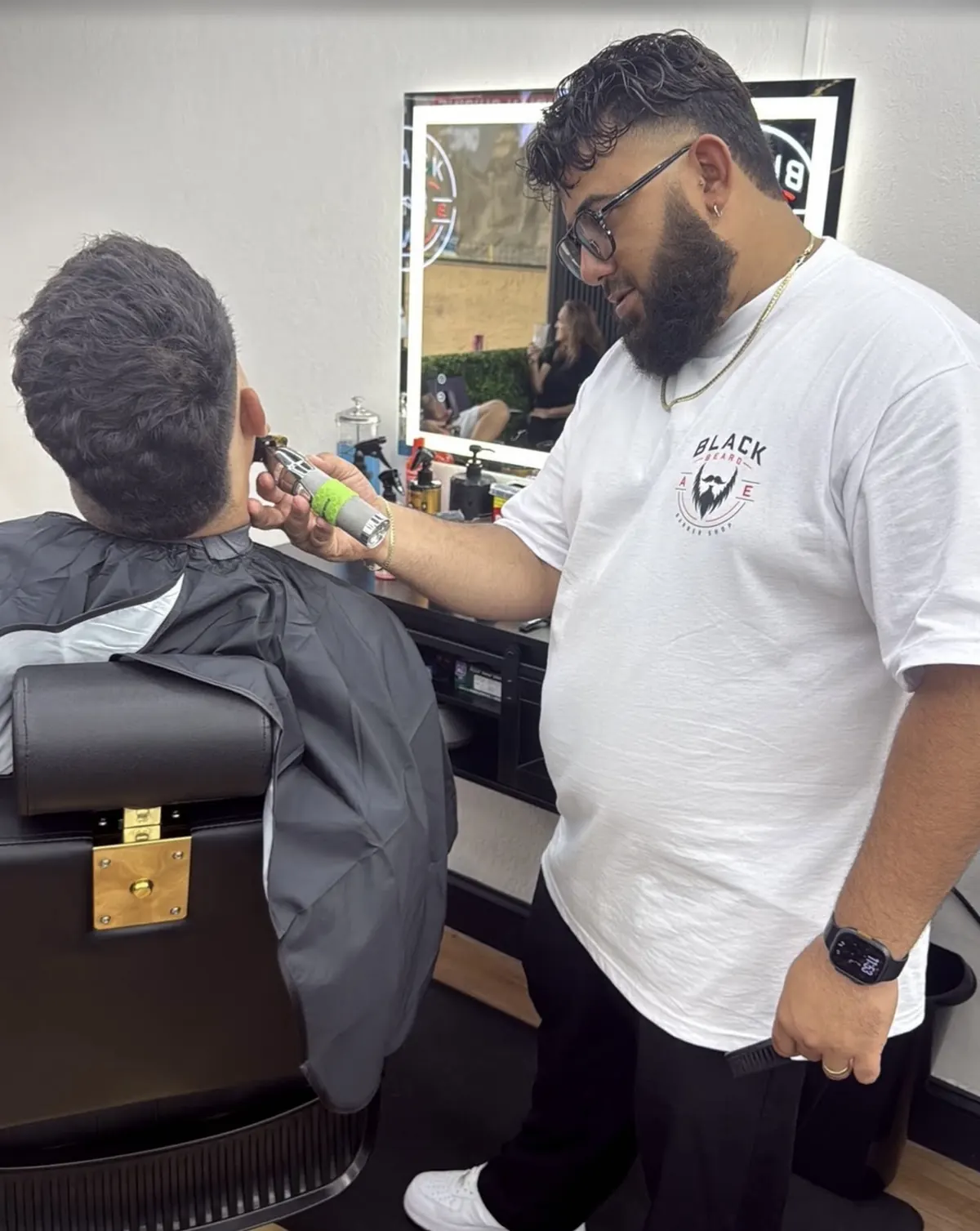 Barber trimming a client's beard at a lit mirror station